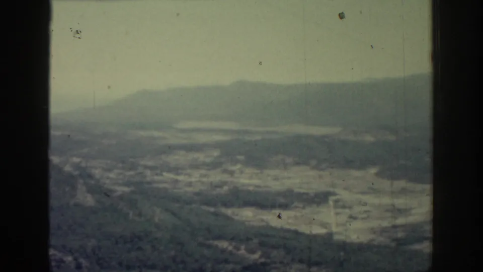 1962 Rhodes, Greece frame showing a wide aerial landscape with dark wooded foreground, open valley areas and distant hazy hills.