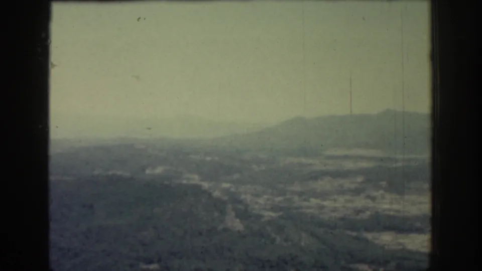 1962 Rhodes, Greece home-movie frame with a broad aerial view of hills, valley settlement traces and hazy mountains beyond.