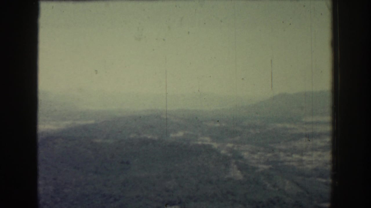1962 Rhodes home-movie frame showing a misty elevated view over hills, distant water, and a washed green-gray sky.