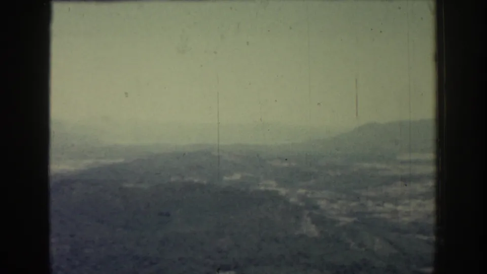 1962 Rhodes home-movie frame showing a misty elevated view over hills, distant water, and a washed green-gray sky.