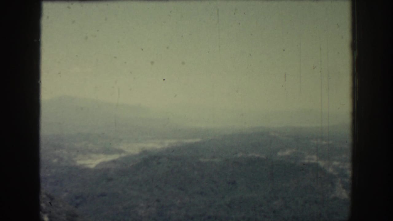 1962 Rhodes landscape frame with a foggy ridgeline, muted hills, and a faint body of water beyond the slopes.