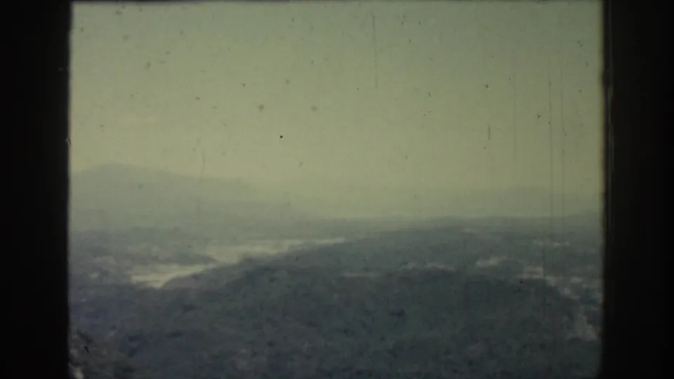 1962 Rhodes landscape frame with a foggy ridgeline, muted hills, and a faint body of water beyond the slopes.