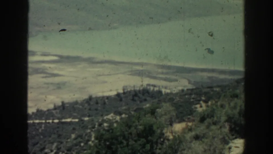 1962 Rhodes frame showing a clearer coastal overlook with water, shoreline, trees, shrubs, and scattered buildings below.