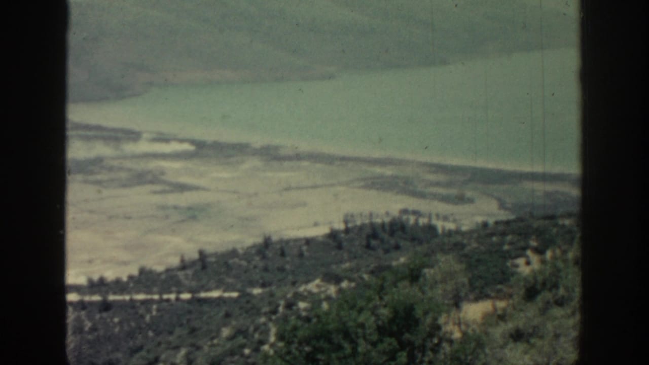 Elevated view of pale shoreline, green hillside, and turquoise water framed by dark film borders.