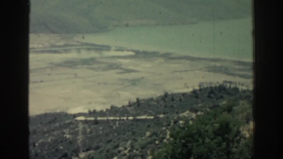 1962 Rhodes travel footage frame with a hillside foreground, scattered trees, light shoreline, and calm blue-green water beyond.