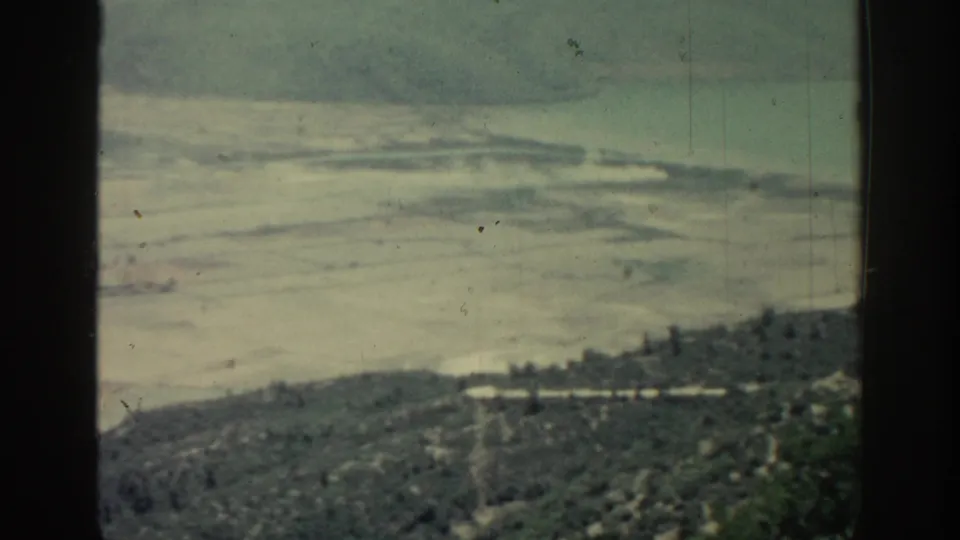 Rhodes 1962 home-movie frame showing a muted coastal panorama with pale waterline, green hillside, and visible 8mm wear.