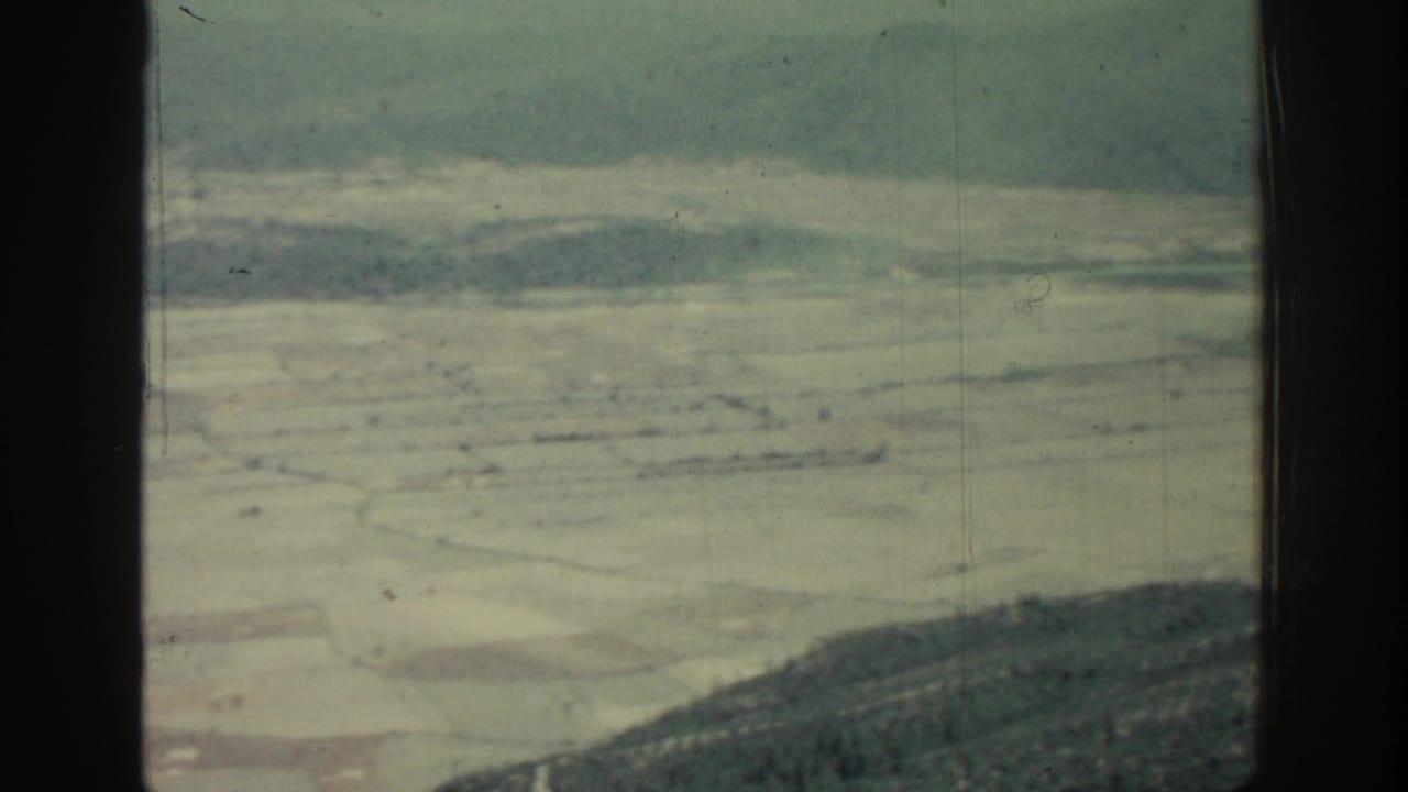 Aerial view of pale fields and wooded hills through dark vertical window bars.