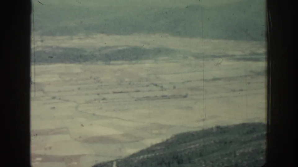 1962 Rhodes home-movie frame showing patchwork fields, hazy hills, and dark window-frame bars from an aircraft-like viewpoint.