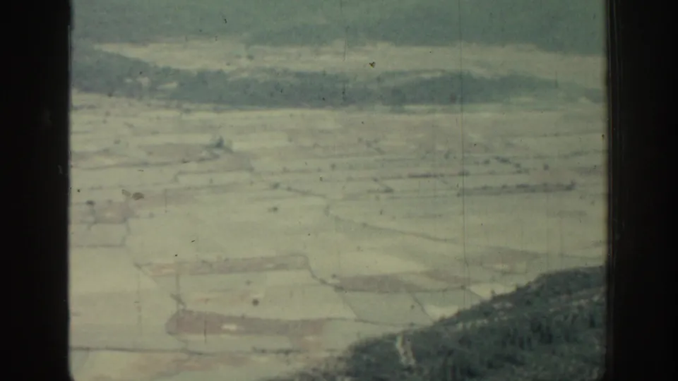 1962 Rhodes aerial view with divided fields, low wooded hills, distant haze, and a dark vertical bar crossing the frame.