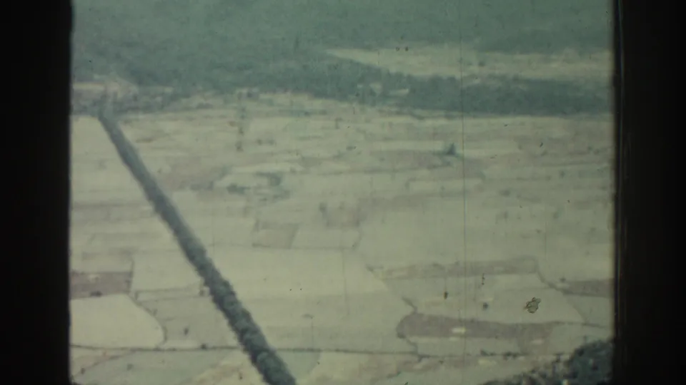 1962 Rhodes travel footage frame showing pale farmland from above with a long dark straight strip crossing the scene.
