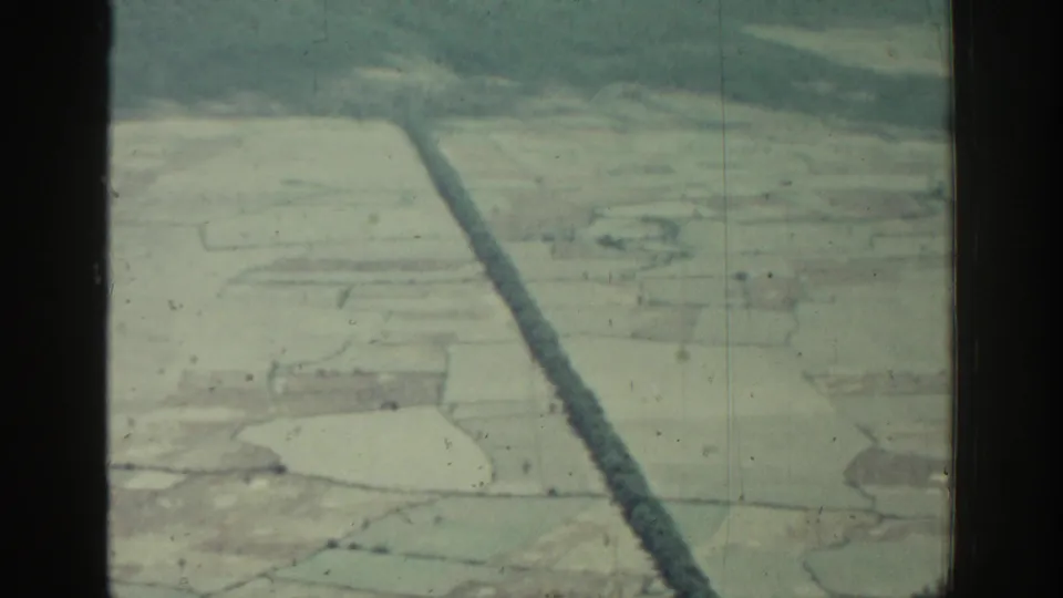 1962 Rhodes aerial home-movie frame with divided rural fields, a straight dark strip, wooded hills, and muted 8mm color.