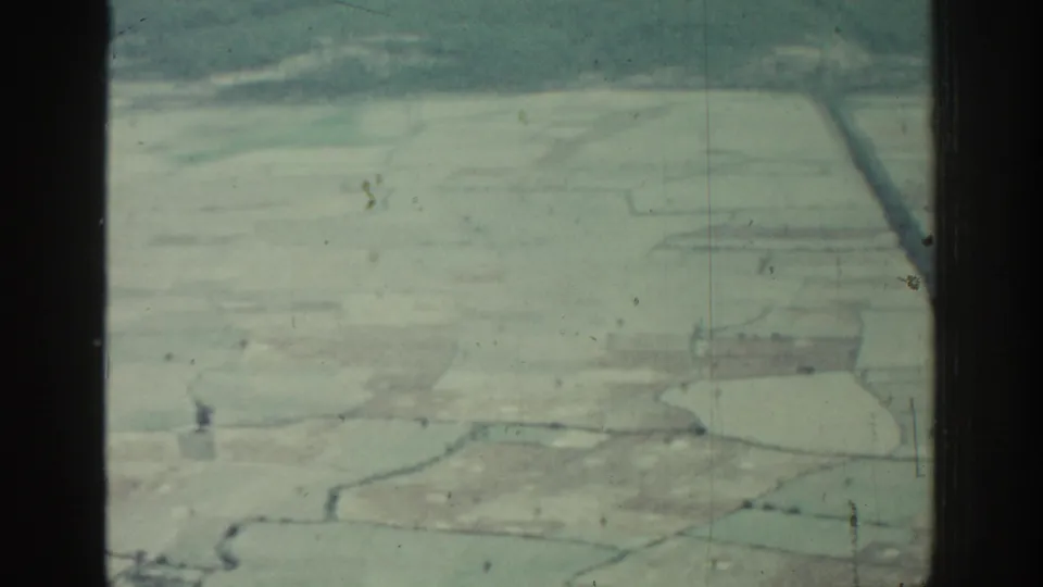 Aerial 8mm frame over Rhodes showing pale rural fields, narrow lines, and dark aircraft window framing.