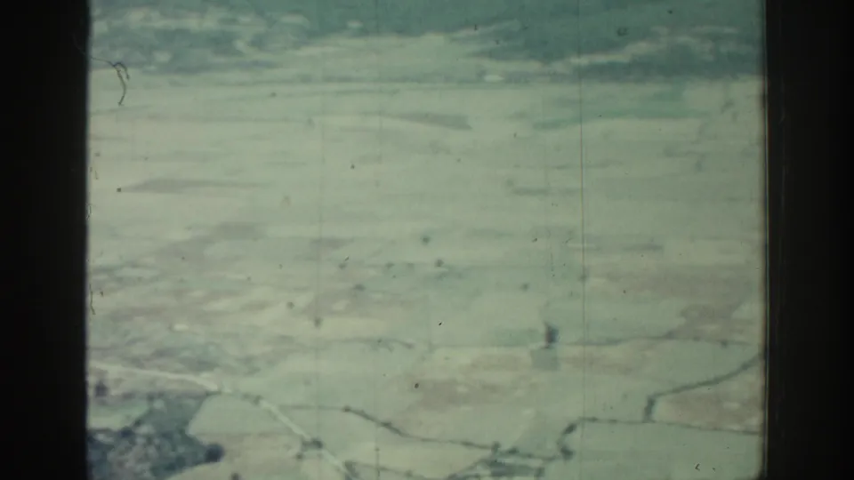 1962 Rhodes aerial footage frame with broad pale fields, window bars, and visible 8mm film wear.