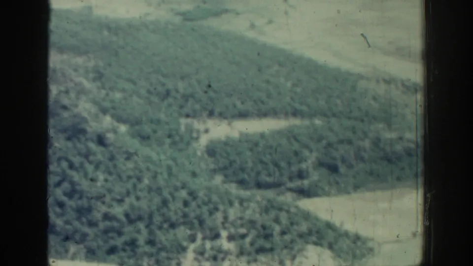 1962 Rhodes home-movie frame showing dense forested ground, light fields, and dark window borders from above.