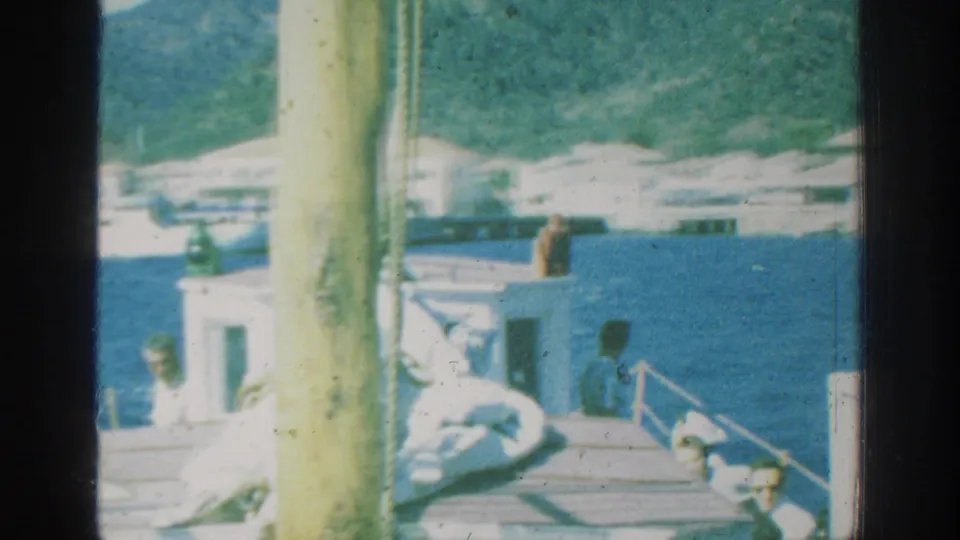 1962 Rhodes frame showing a boat deck on blue water, distant people, a white structure, and a mountainous coastline.
