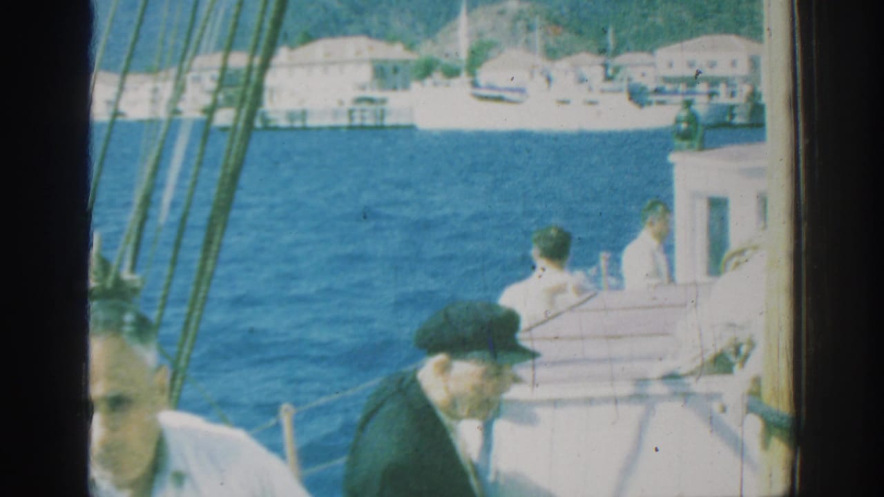Men on a sailboat deck with blue water and shoreline buildings behind them.