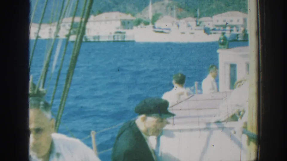 1962 Rhodes, Greece home-movie frame showing adult men on a sailboat deck, with rigging, blue sea, and pale waterfront buildings in the background.