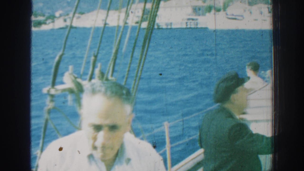 A man in a white shirt faces the camera on a sailboat with another man at the rail.