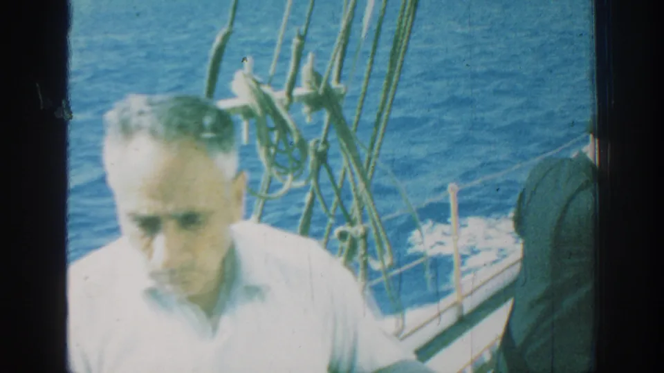 1962 Rhodes home-movie frame with a close adult passenger in a white shirt, sailboat rigging, deck rail, and textured blue water behind.