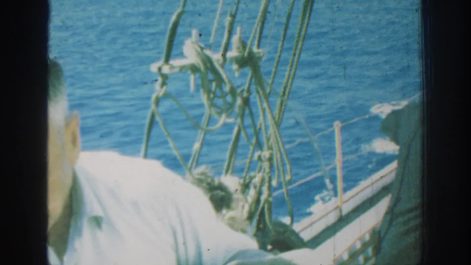A cropped 1962 Rhodes sailboat frame shows an adult passenger near the camera, with rigging, railings, wake, and blue sea visible around him.