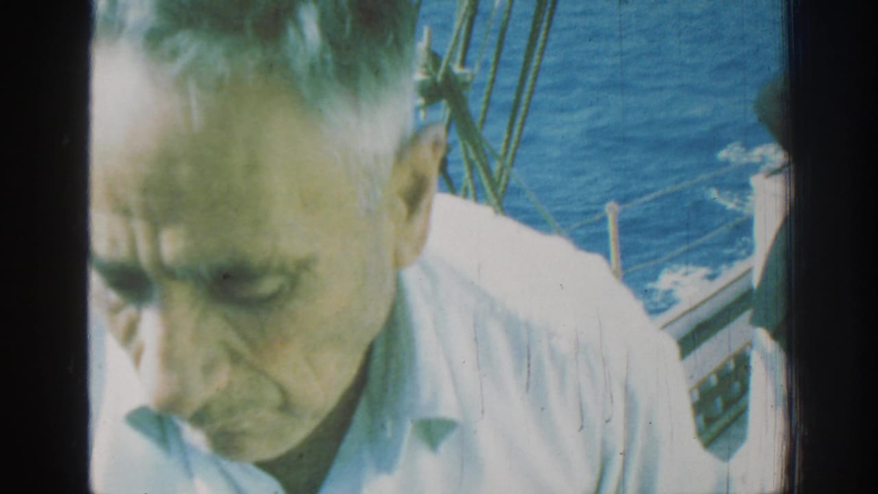 1962 Rhodes home-movie frame showing a man in a light shirt near the camera aboard a moving boat, with rigging and blue sea behind him.