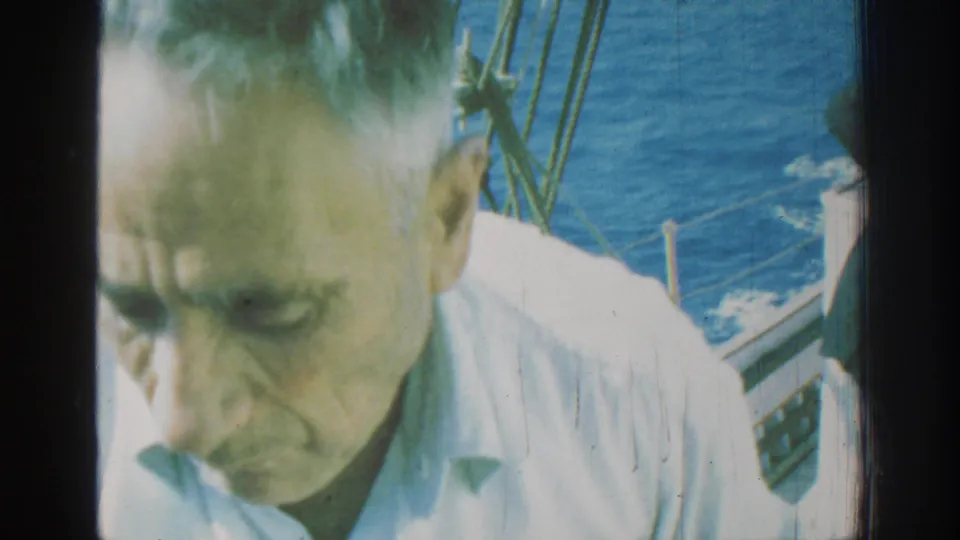 1962 Rhodes home-movie frame showing a man in a light shirt near the camera aboard a moving boat, with rigging and blue sea behind him.