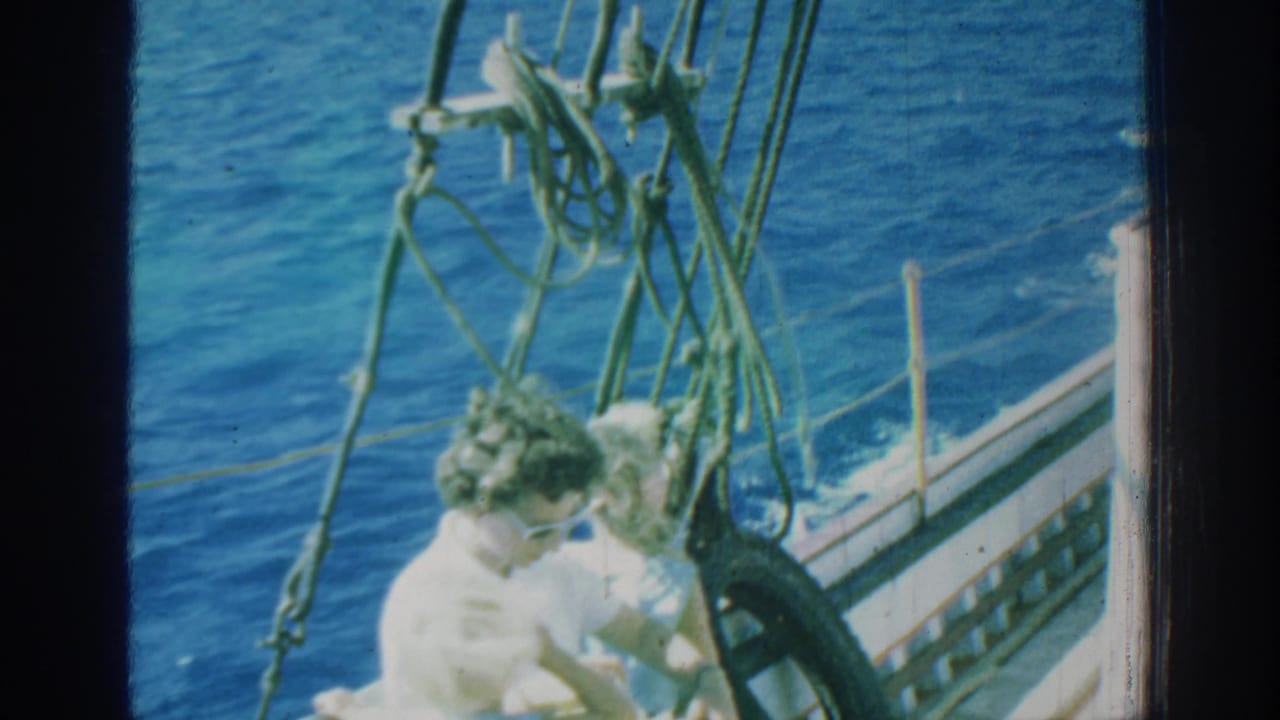 1962 Rhodes frame of a woman in sunglasses seated near another passenger on the bow of a moving sailboat, with green rigging over blue water.