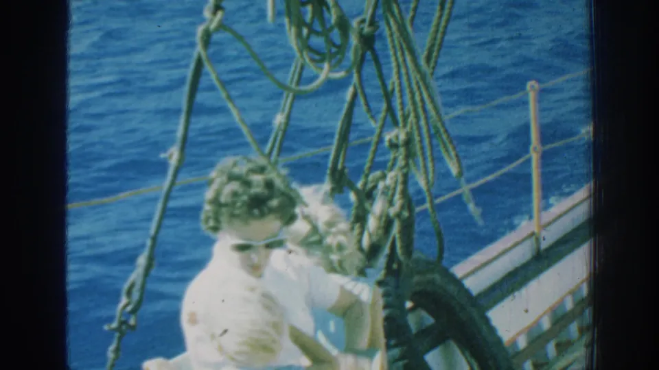 1962 Rhodes home-movie frame showing a woman in sunglasses and another passenger seated on the bow near ropes, railing, and a tire fender.