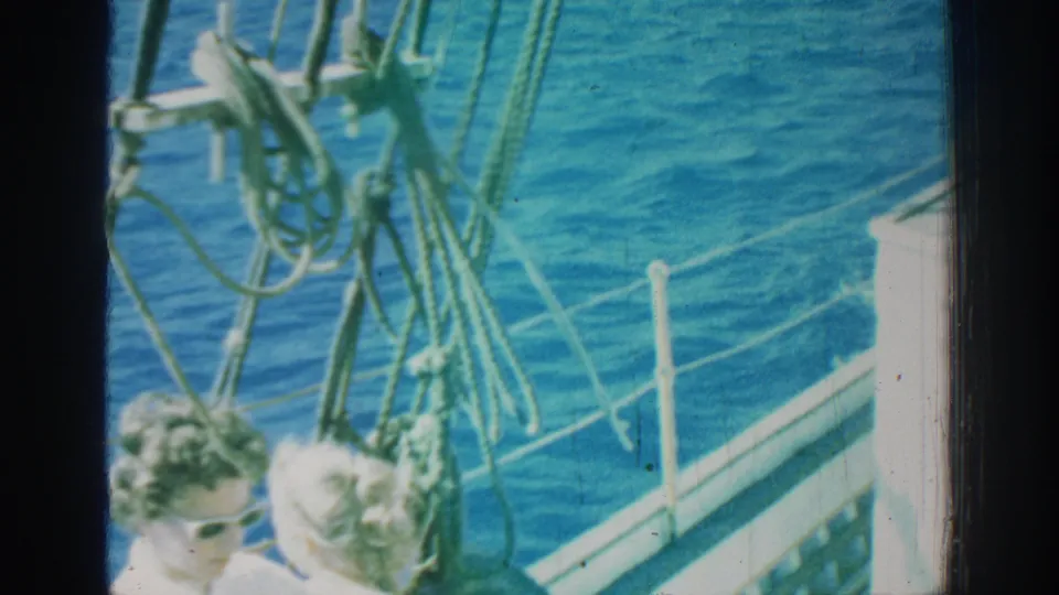 1962 Rhodes frame with passengers partly seen beneath green rigging on a sailboat bow, surrounded by railing, rope, fender, and sunlit sea.