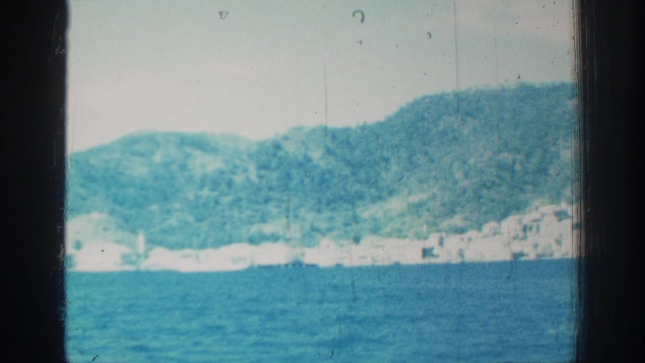 Coastal village across blue water with green hills behind it and dark film borders.
