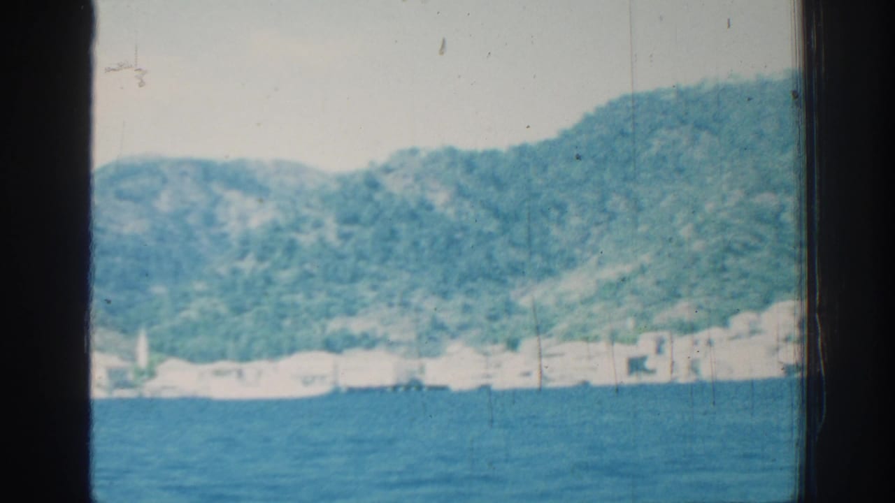 White coastal buildings and small masts along the shore below green hills.