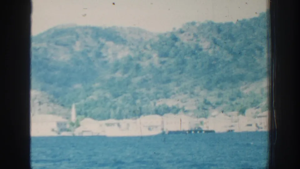 Soft 8mm frame from Rhodes showing choppy blue water, a bright shoreline, and a green hillside rising behind it.