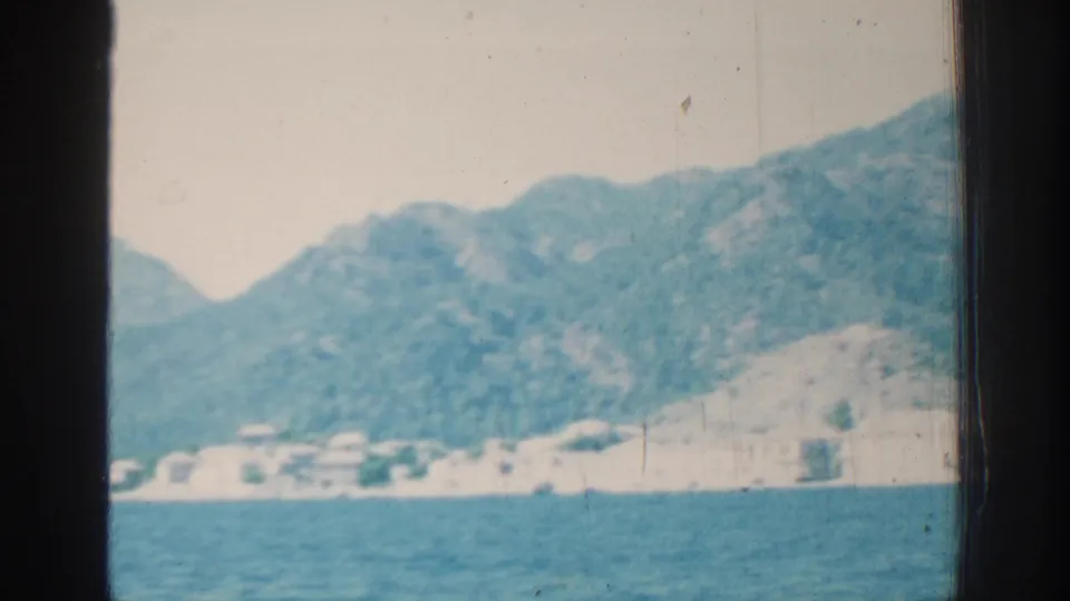 Rhodes, Greece shoreline seen from water in 1962 footage, with low white buildings and green mountains under a pale sky.