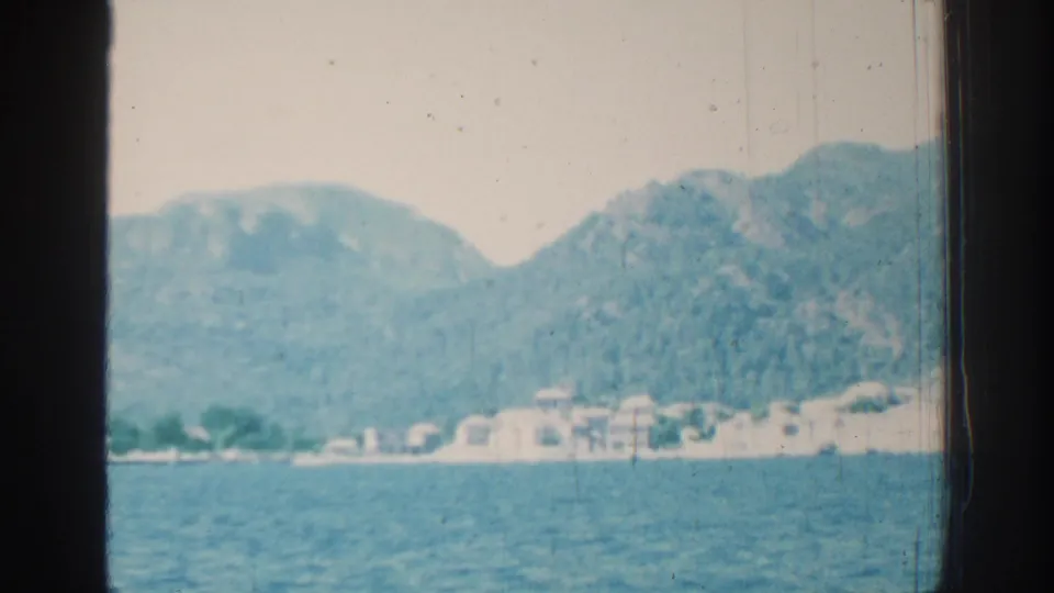 A soft 8mm frame of the Rhodes coastline, showing rippled blue water, pale buildings, and steep green hills in the background.