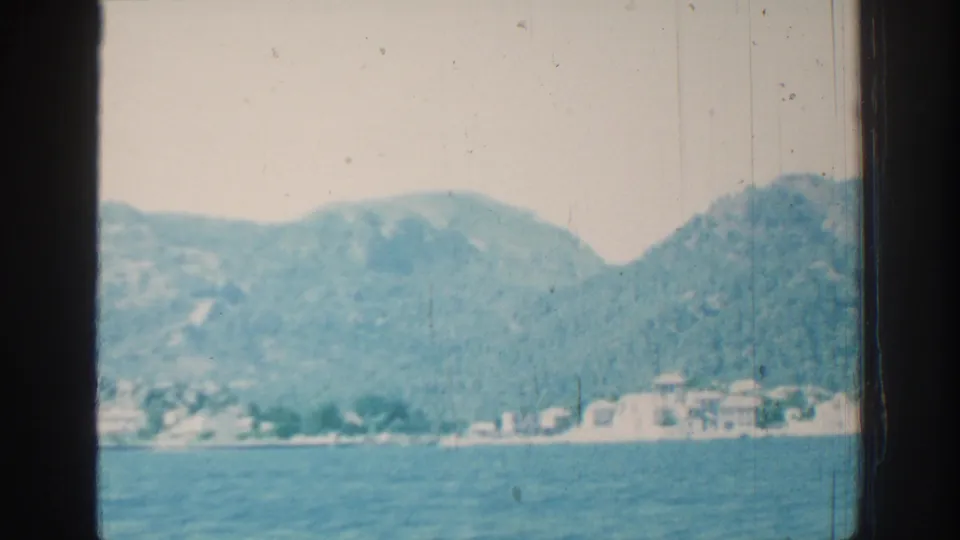 Late frame from the 1962 Rhodes coastal shot, with blue water, white waterfront structures, and forested mountain slopes beyond.