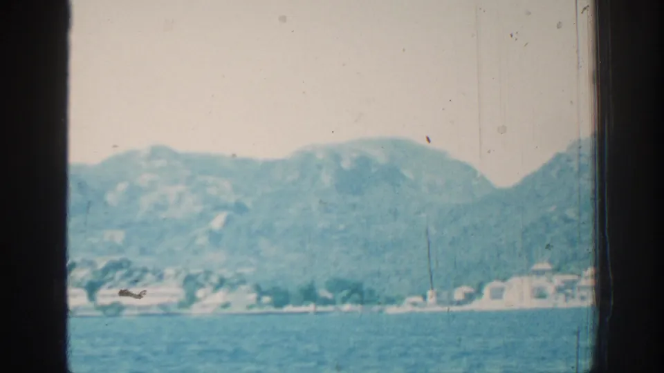 1962 Rhodes coastal view from a moving vessel, with blue water, hazy hills, and distant pale shoreline buildings framed by dark window posts.