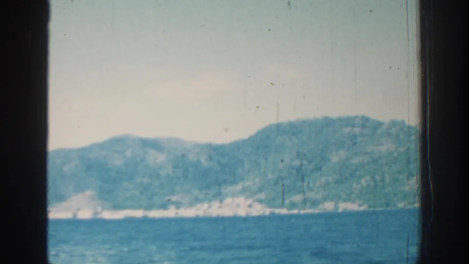 Rhodes coastline appears beyond a dark window divider, with rippled turquoise water, hazy sky, and pale settlement along the shore.