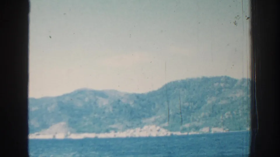 A visitor-shot 8mm frame shows the Rhodes shore from offshore, with low ripples, green hills, pale rock, and a dark window edge.