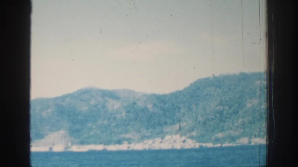 The final frame holds on the Rhodes coastline, showing blue water, hazy green hills, pale shore structures, and a dark vessel window border.