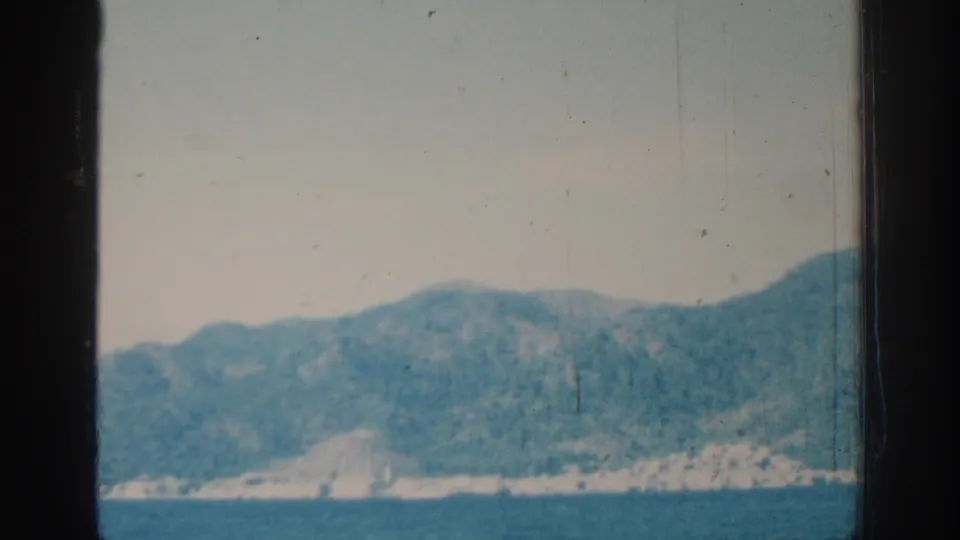 1962 Rhodes coastal view across blue sea, with tree-covered hills and shoreline softened by 8mm film texture.