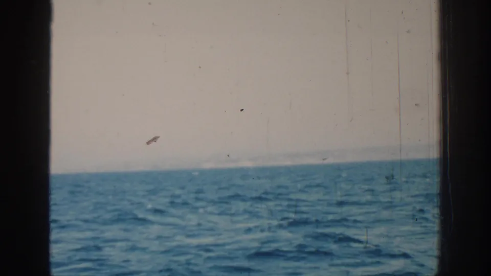 1962 home-movie frame of the sea near Rhodes, with a low hazy horizon, blue water, dark boat edge, and aged 8mm texture.