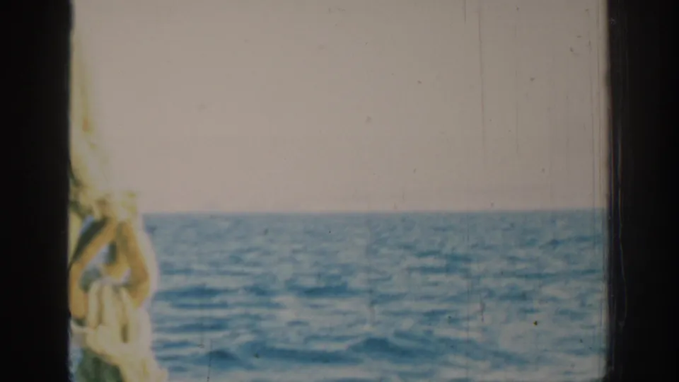 1962 Rhodes boat-view frame showing blue waves, pale sky, dark vertical framing, and a blurred rope or line near the center.