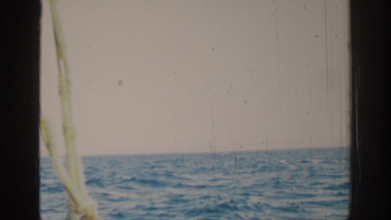 Open sea and pale sky seen past dark film edges and sailboat rigging.