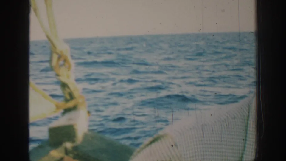 1962 Rhodes home-movie frame from a sailboat, showing deck texture, yellow sail rigging, blue water, and a bright washed sky.