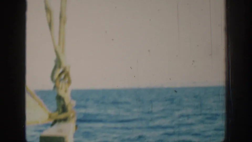 1962 Rhodes sailing footage frame with yellow sail rigging in the foreground and the blue sea stretching to a low horizon.