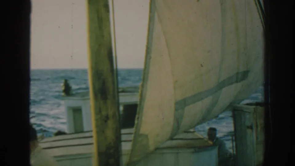 1962 Rhodes 8mm frame of a small sailing boat on blue water with mast, cabin, sail, and a person on deck.