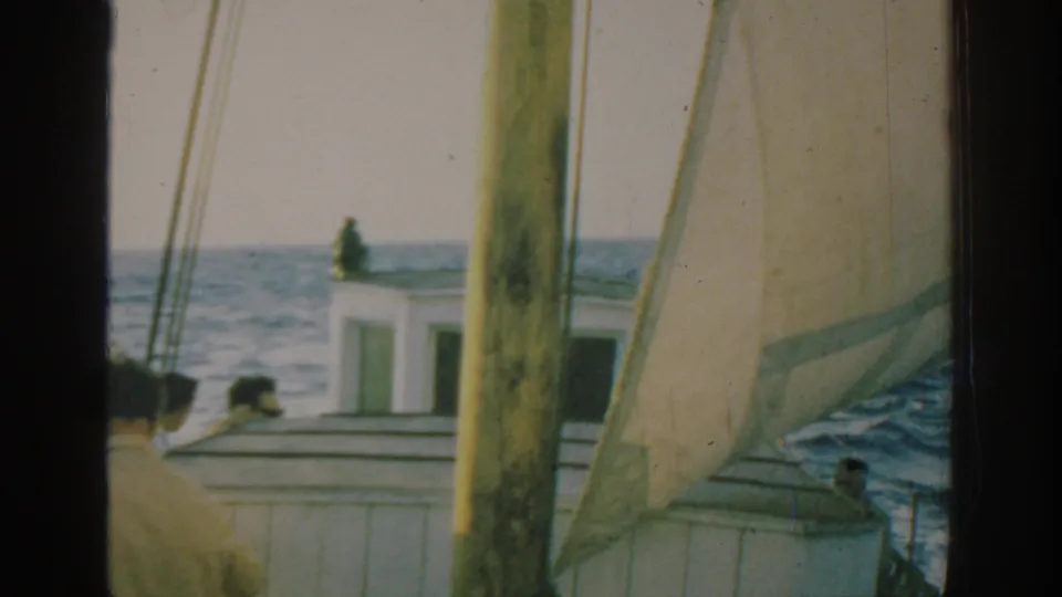 1962 Rhodes 8mm frame with a white sail, mast, cabin roof, blue sea, and people aboard a small boat.