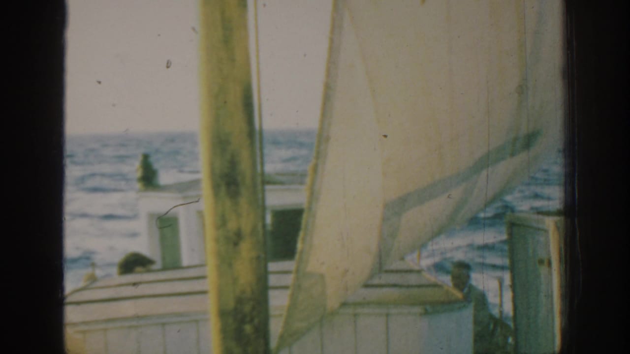 White sailboat with pale sail and passengers on blue water, partly framed by dark vertical edges.