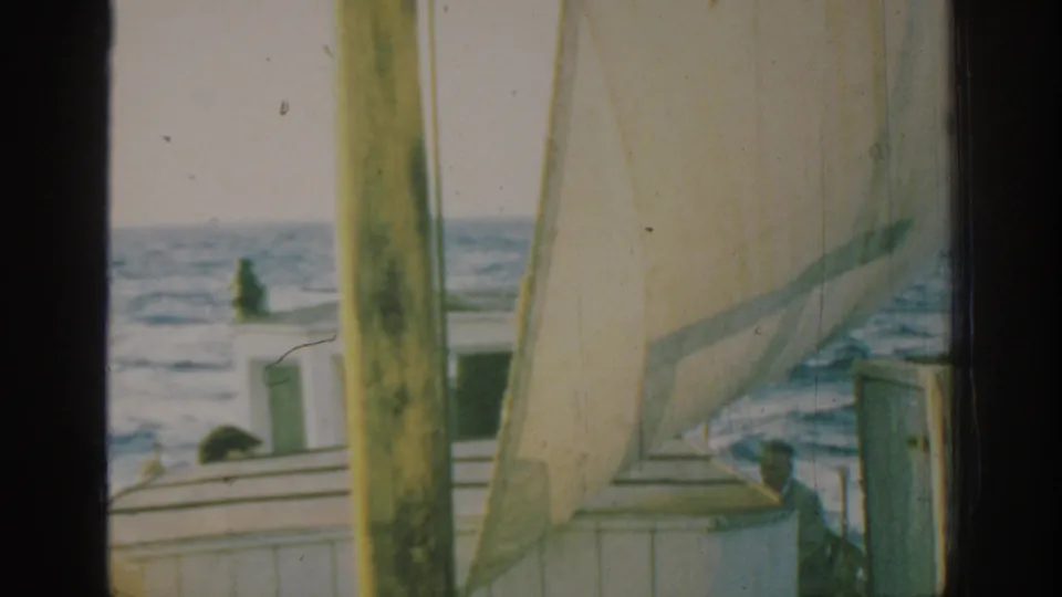 1962 Rhodes 8mm view of a white sailboat with a pale sail and passengers on deck, seen through a dark window-like frame.