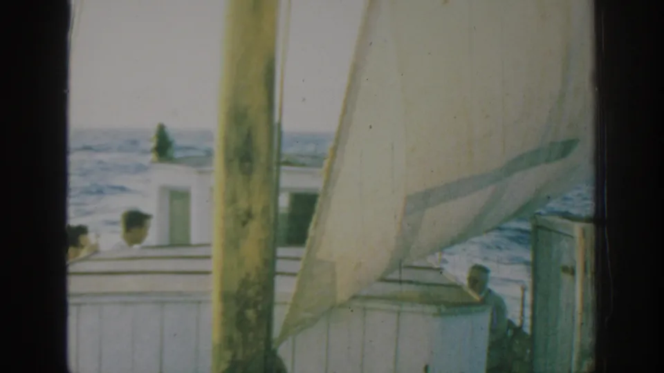 Home-movie frame of a sailboat off Rhodes with passengers on deck, pale sail fabric, mast, and sea visible beyond the cabin.